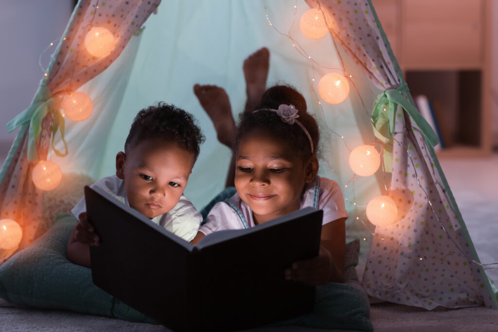 Two Black children reading a book under a fort made of bedsheets with small lights.